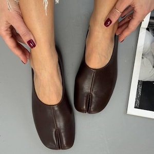 May include: A pair of dark brown leather split-toe flats. The shoes have a rounded toe and a split design separating the big toe from the other toes. The flats are a solid color and appear to be made of smooth leather.