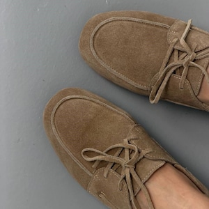 May include: A pair of light brown suede moccasin-style shoes with laces. The shoes have a rounded toe and a stitched design around the top. The laces are tied in a bow. The shoes are on a gray surface.