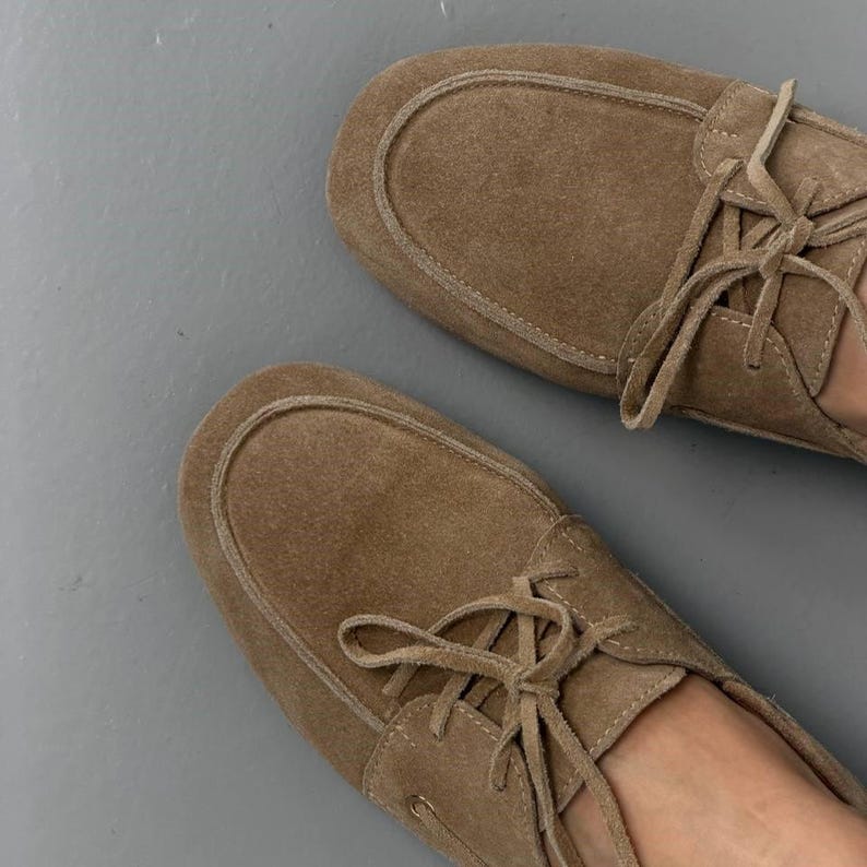 May include: A pair of light brown suede moccasin-style shoes with laces. The shoes have a rounded toe and a stitched design around the top. The laces are tied in a bow. The shoes are on a gray surface.