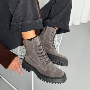 May include: Gray suede ankle boots with black laces and a chunky black sole. The boots have a side zipper and are worn with dark gray denim pants. The boots are a neutral color and can be worn with many outfits.