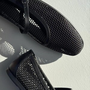 May include: A pair of black mesh ballet flats featuring a rounded toe, a thin adjustable strap with a small metal buckle across the top, and solid black trim along the edges. The shoes are constructed from a semi-sheer, grid-patterned mesh material.