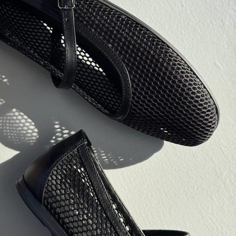 May include: A pair of black mesh ballet flats featuring a rounded toe, a thin adjustable strap with a small metal buckle across the top, and solid black trim along the edges. The shoes are constructed from a semi-sheer, grid-patterned mesh material.