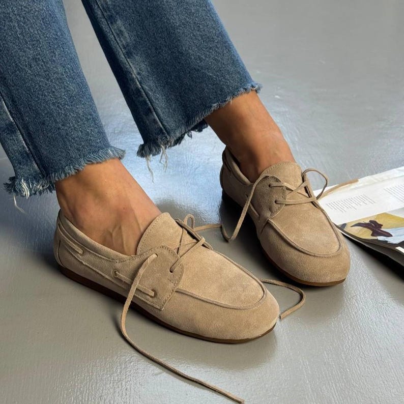 May include: Tan suede boat shoes with laces. The shoes have a rounded toe and a stitched sole. The image also shows blue jeans with frayed hems.