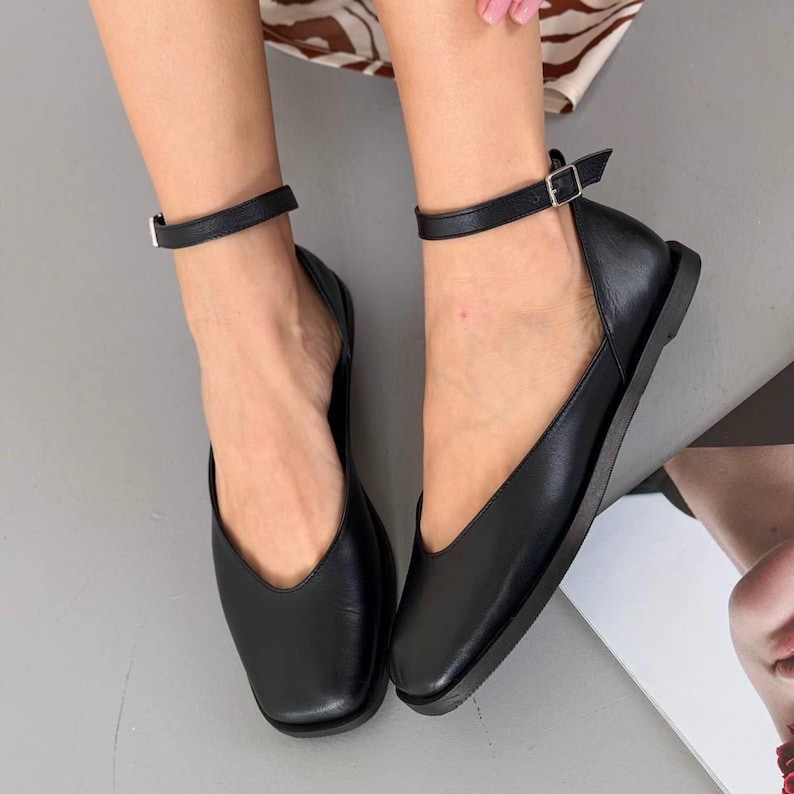 May include: Black leather flats with ankle straps. The shoes have a square toe design and a small buckle closure on the ankle strap. The flats are on a gray surface.
