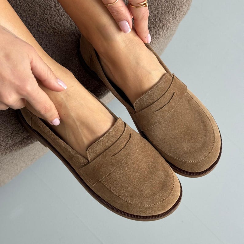 May include: A pair of tan suede penny loafers featuring a rounded toe, a classic strap across the vamp, and a low-profile design. The shoes have a smooth, matte texture and a dark brown sole, showcasing a clean, slip-on construction for everyday wear.