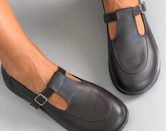 May include: A pair of dark brown leather T-strap shoes featuring a rounded toe, a smooth finish, and a thin adjustable strap across the instep secured with a small silver-toned metal buckle. The shoes have a low-profile design and a flat sole.