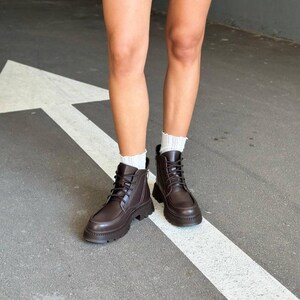 May include: Brown leather boots with a lace-up design and a chunky sole. The boots are worn with white socks. The image shows the lower legs and feet of a person standing on a gray surface with a white arrow.