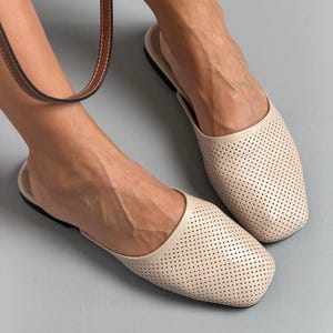 May include: A pair of beige slip-on mules featuring a square toe design and a perforated upper surface. The shoes have a flat black sole and a smooth, solid-colored trim around the opening, providing a clean and simple aesthetic for footwear.