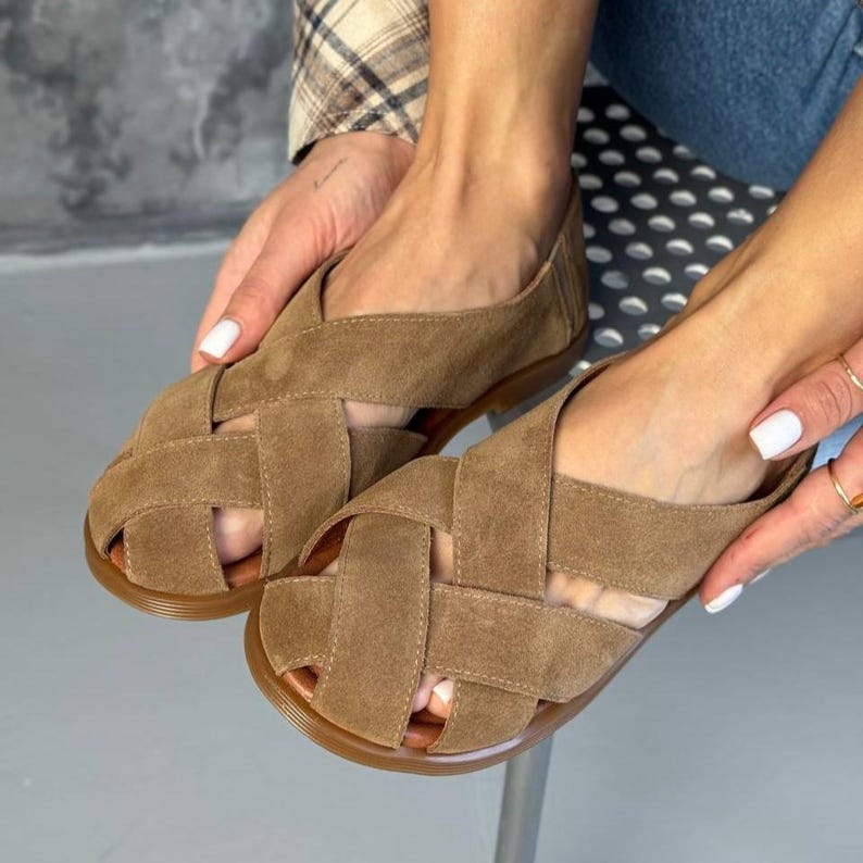 May include: Brown suede sandals with a woven design. The sandals have a flat sole and a crisscross pattern across the top. The shoes are a light brown color and appear to be made of soft suede material. The sandals are a casual style.