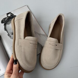 May include: A pair of beige suede penny loafers featuring a rounded toe, a classic strap across the vamp with a cutout detail, and a flat brown sole. The shoes have a soft, matte texture and a neutral light tan color throughout the exterior.