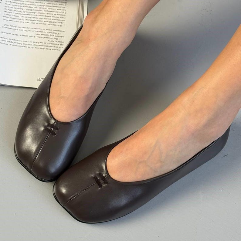 Op de afbeelding: Een paar donkerbruine leren ballerina's met een vierkante neus en een klein decoratief stikseldetail op de wreef. De schoenen hebben een laag profiel en een eenvoudig, elegant ontwerp, geschikt voor dagelijks gebruik.
