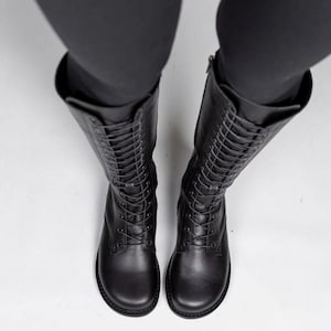 May include: A pair of black mid-calf boots featuring a rounded toe, a full-length lace-up front with matching black laces, and a side zip closure. The boots have a smooth finish and a flat sole, extending up the leg to the mid-calf area.