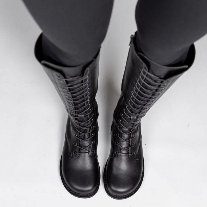 May include: A pair of black mid-calf boots featuring a rounded toe, a full-length lace-up front with matching black laces, and a side zip closure. The boots have a smooth finish and a flat sole, extending up the leg to the mid-calf area.