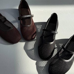 May include: Four Mary Jane style shoes featuring a mesh upper construction. Two shoes are dark brown and two are black. Each shoe has a thin strap across the instep with a buckle closure and a rounded toe design, showcasing a textured grid pattern.