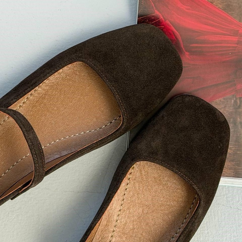 May include: A pair of dark brown suede flats with a rounded toe and a strap across the top of the foot. The interior is a lighter brown color, and the shoes appear to be unworn. The shoes are on a light gray surface.