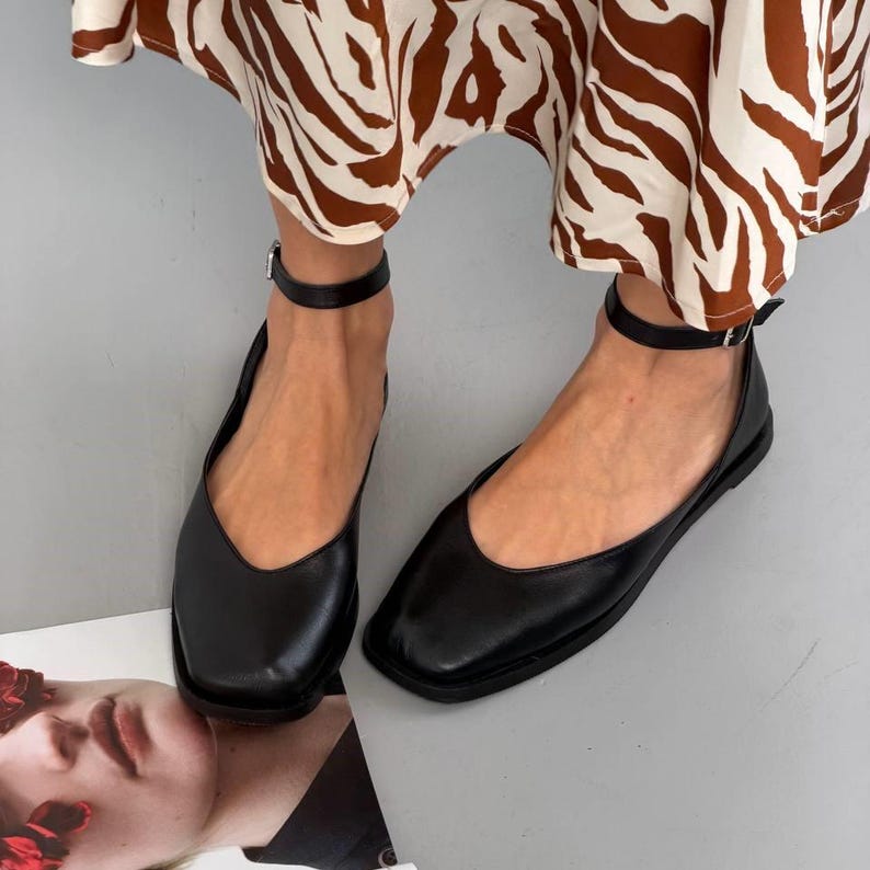 May include: Black leather flats with a square toe and ankle strap. The shoes have a low heel and a simple, elegant design. The ankle straps fasten with a small buckle. The flats are shown with a patterned skirt.