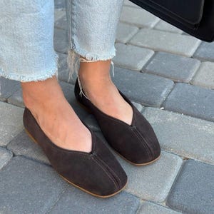 Pode incluir: Sapatilhas de camur&ccedil;a castanho-escuro com bico quadrado e decote em V. Os sapatos t&ecirc;m uma costura costurada no centro e uma sola castanho-claro. Usadas com jeans de ganga azul claro.
