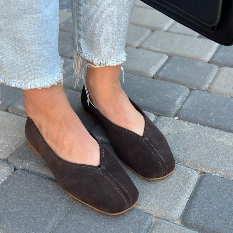 Pode incluir: Sapatilhas de camur&ccedil;a castanho-escuro com bico quadrado e decote em V. Os sapatos t&ecirc;m uma costura costurada no centro e uma sola castanho-claro. Usadas com jeans de ganga azul claro.