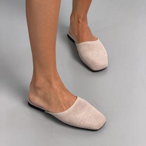 May include: A pair of light beige slip-on mules featuring a perforated texture across the upper part of the shoe. These flat shoes have a square toe design and a thin black sole, providing a simple and functional footwear option for daily use.