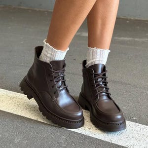 May include: Brown leather ankle boots with a lace-up design and a chunky sole. The boots have a rounded toe and a zipper on the side. The boots are worn with white, patterned socks.