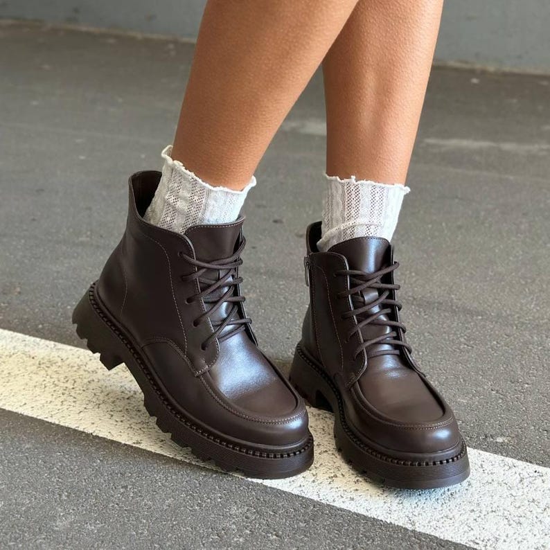 May include: Brown leather ankle boots with a lace-up design and a chunky sole. The boots have a rounded toe and a zipper on the side. The boots are worn with white, patterned socks.