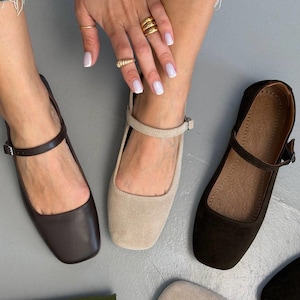 May include: Three pairs of women's flat shoes in brown, beige, and black. Each shoe has a square toe and a strap that buckles around the ankle. The shoes are displayed on a gray surface.