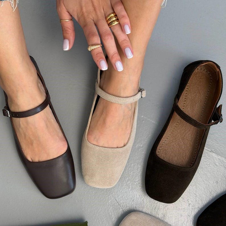 May include: Three pairs of women's flat shoes in brown, beige, and black. Each shoe has a square toe and a strap that buckles around the ankle. The shoes are displayed on a gray surface.