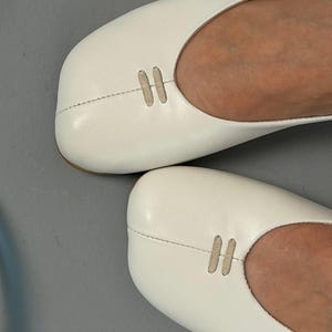 Op de afbeelding: Close-up van twee witte leren ballerina's. Elke schoen heeft een decoratief, gestikt ontwerp aan de bovenkant. De schoenen zijn effen wit van kleur en lijken ongedragen.