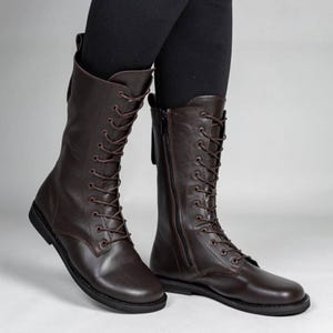 Puede incluir: Un par de botas de media caña en color marrón oscuro con cordones frontales a juego y ojales metálicos. Las botas presentan un acabado liso, puntera redondeada, suela plana negra y una cremallera lateral para facilitar su cierre.