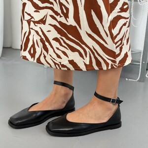 May include: Black leather flats with ankle straps. These shoes have a square toe design and a small buckle closure on the ankle strap. The flats are shown with a brown and white patterned skirt.
