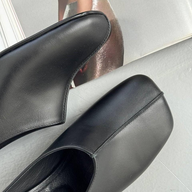 May include: Close-up of two black leather shoes. The shoes have a square toe design and a low heel. The leather appears smooth and the stitching is visible along the edges.
