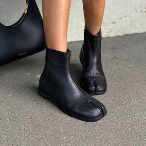May include: Black leather ankle boots with a split toe design. The boots have a low heel and a zipper on the back. A black leather handbag is visible in the background.