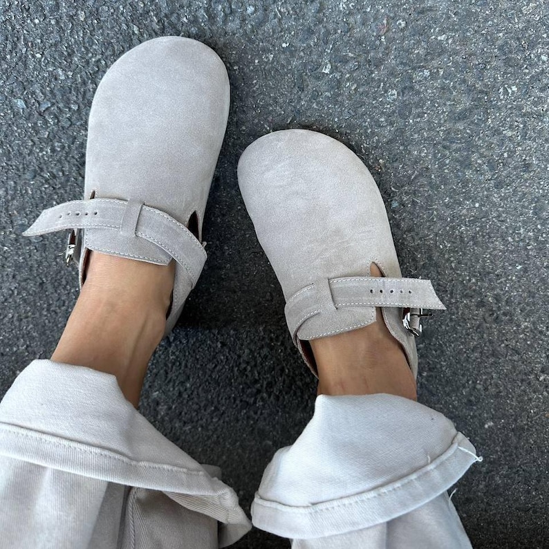 May include: A pair of light gray suede clogs with adjustable ankle straps and silver buckles. The clogs have a rounded toe and a minimalist design, suitable for casual wear. The shoes are shown from a top-down perspective.