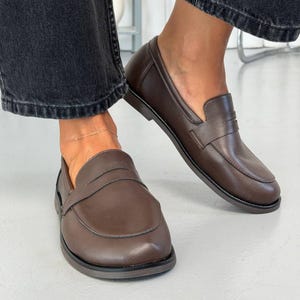 May include: A pair of dark brown leather loafers featuring a classic penny slot design across the vamp, a rounded toe, and a low heel. The shoes have a smooth, matte finish and are worn with dark grey denim jeans, showcasing a clean silhouette.