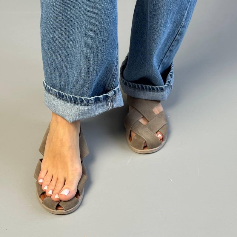 May include: Close-up of gray sandals with a woven design. The sandals have a light brown sole and are worn with blue jeans. The jeans are cuffed at the ankles, revealing the sandals.