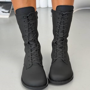 Könnte beinhalten: Ein Paar wadenhohe Stiefel in Mattschwarz mit abgerundeter Zehenpartie, durchgehender Schnürung vorne mit passenden schwarzen Schnürsenkeln und einem seitlichen Reißverschluss. Die Stiefel haben eine glatte Oberfläche und eine robuste Sohle.