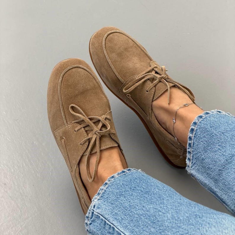 May include: A pair of light brown suede boat shoes with matching laces. The shoes have a rounded toe and a stitched design around the top. The shoes are worn with blue jeans.