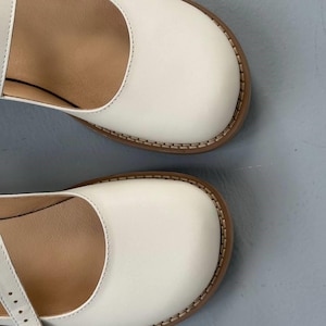 May include: Close-up of two white Mary Jane style shoes. The shoes have a rounded toe, a strap, and a brown sole with visible stitching. The interior of the shoe is a light brown color.