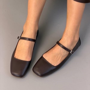 May include: A pair of dark brown leather Mary Jane flats. The shoes have a square toe design and a thin strap that buckles across the top of the foot. The flats are displayed on a gray surface.