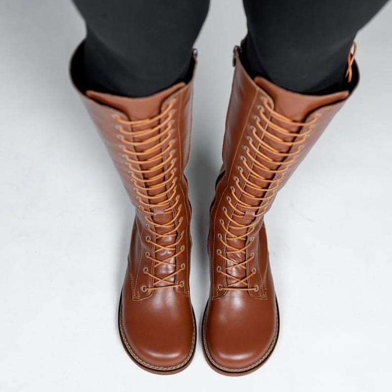 May include: A pair of knee-high brown leather boots featuring a rounded toe, a full-length lace-up front with matching brown laces, and metal eyelets. The boots extend to the mid-calf and are worn with black leggings, showing a smooth finish.