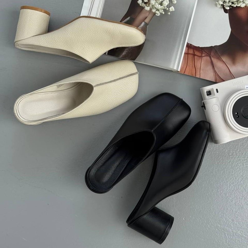 May include: Two pairs of leather mules in cream and black. The cream-colored mules have a block heel and a rounded toe. The black mules have a similar block heel and a square toe. The shoes are displayed on a gray surface.