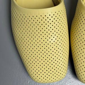 May include: A pair of pale yellow slip-on shoes featuring a perforated design across the entire upper surface. The shoes have a rounded square toe shape and a smooth, solid-colored trim along the opening, showcasing a consistent dotted pattern.