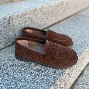 May include: A pair of brown suede loafers with a classic design. The shoes have a rounded toe, a strap across the vamp, and a dark brown sole. The loafers are displayed on a textured gray surface.