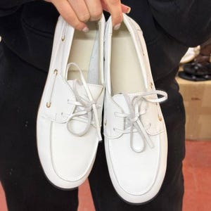 May include: A pair of white boat shoes with light-colored laces. The shoes have a classic design with a rounded toe and a low profile. The shoes are made of a smooth, white material and feature gold-colored eyelets.