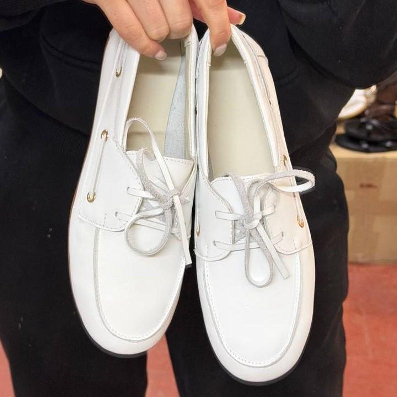 May include: A pair of white boat shoes with light-colored laces. The shoes have a classic design with a rounded toe and a low profile. The shoes are made of a smooth, white material and feature gold-colored eyelets.