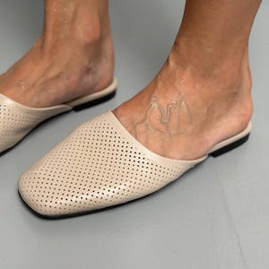 May include: A pair of beige slip-on mules featuring a square toe design and a perforated upper surface. The footwear has a flat black sole and a smooth, light-colored finish across the front, providing a simple and functional style for daily wear.