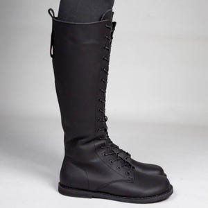 May include: A knee-high black boot featuring a full-length lace-up front closure with matching black laces and eyelets. The boot has a smooth matte finish, a rounded toe, a flat sole, and a vertical pull tab at the top rear of the shaft.