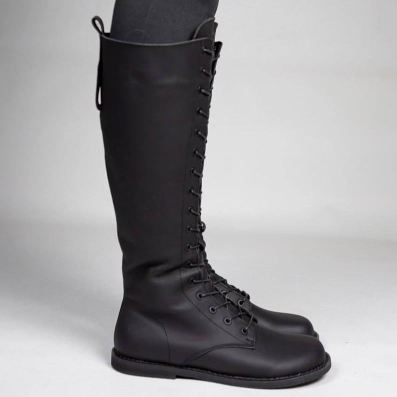 May include: A knee-high black boot featuring a full-length lace-up front closure with matching black laces and eyelets. The boot has a smooth matte finish, a rounded toe, a flat sole, and a vertical pull tab at the top rear of the shaft.