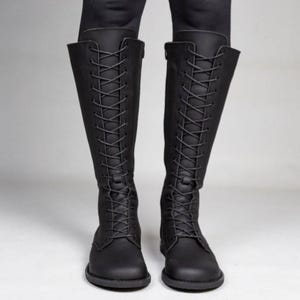 May include: A pair of knee-high black boots featuring a full-length front lace-up closure with matching black laces and eyelets. The boots have a rounded toe, a flat sole, and a smooth matte finish, extending up to the lower calf area.