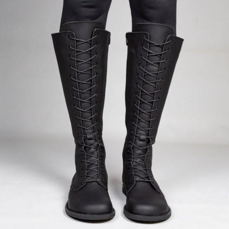 May include: A pair of knee-high black boots featuring a full-length front lace-up closure with matching black laces and eyelets. The boots have a rounded toe, a flat sole, and a smooth matte finish, extending up to the lower calf area.